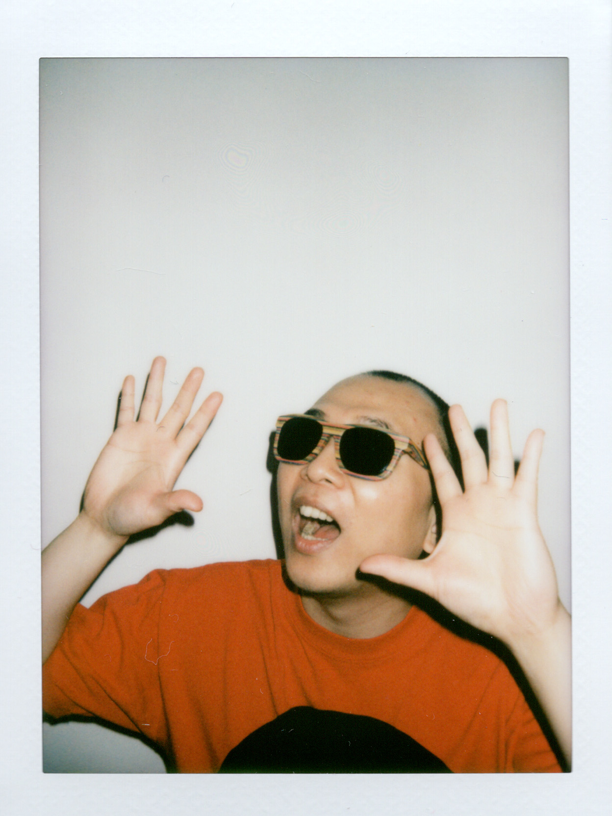 Mike Mai is wearing sunglasses with a colorful frame and a red t-shirt. He has both hands raised up toward the camera with their palms facing outward, fingers spread wide, as if he is being startled by a monster off screen but in a playful way. Their mouth is open wide saying AHHHH!. The visual has a classic film camera aesthetic with slightly muted, warm tones and soft lighting. The background is a simple off-white wall, which makes Mike and his vibrant red shirt really pop. The whole vibe is super spontaneous and fun, like someone caught him in the middle of goofing around.
