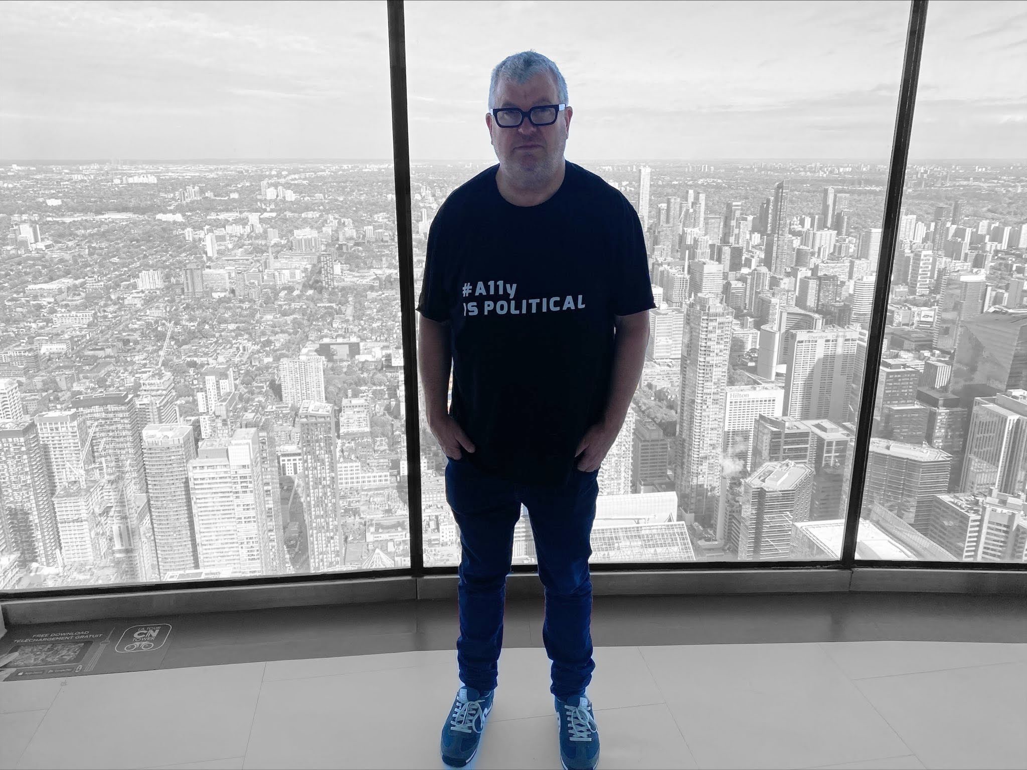 Steve wearing an #a11y is political t-shirt. a view of Toronto from the top of the CN tower in the background.
