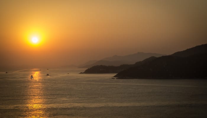 A deep orange sun setting, with a hazy sky. There are a few boats in the water.