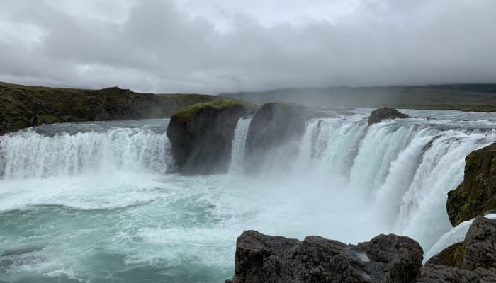 A curved waterfall split into three. The sky is grey with a thick cloud.