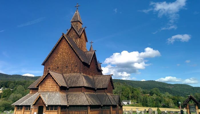 A fully wooden Stave church and graveyard. The sky is mostly clear and blue, with a tree covered hill in the background.