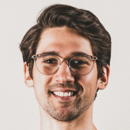 A person with short, dark hair and glasses smiles at the camera against a light background.