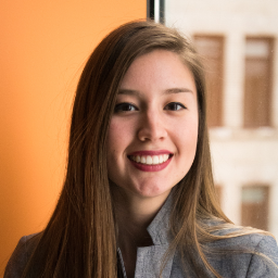 A person with long brown hair smiles in front of a window. The background is a combination of an orange wall and an out-of-focus building exterior. The person is wearing a gray jacket.