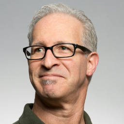 An older man with short gray hair and glasses, wearing a dark shirt, looks upwards and slightly to the side against a plain gray background.