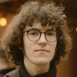 A person with curly hair and glasses looks at the camera. They are wearing a dark turtleneck sweater. The background is softly blurred with warm, glowing lights.