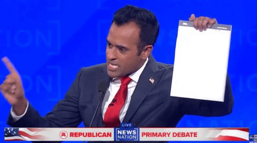 From a screen labeled Republican Primary Debate, a man of Indian heritage wearing a blue suit and red tie wags has finger to someone off camera while holding a white legal notepad in his other hand with the following text scrawled on it: