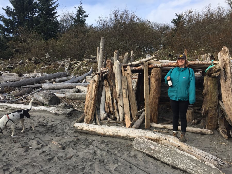 A woman standing in front of a driftwood structure holding a cider bottle.