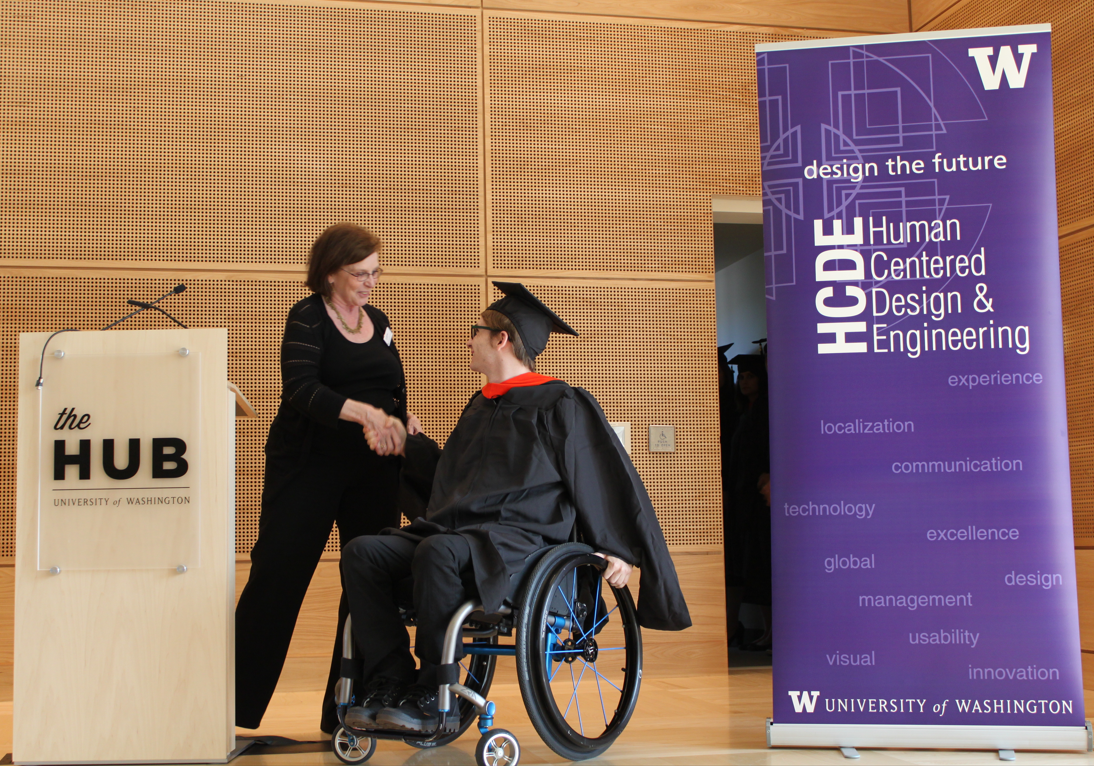 Luke receiving his UW M.S. grad diploma on a stage. Luke is in a manual wheelchair.