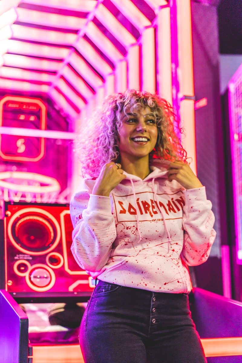 a smiling person in a pink hoodie, standing in front of a bright pink lighted arcade basketball game. 